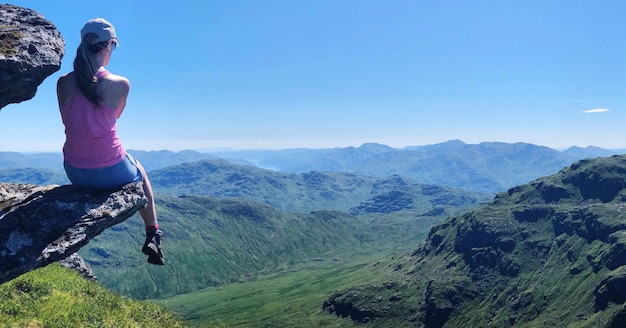 Eine Frau in voller Länge sitzt auf einem Felsen gegen Berge und Himmel