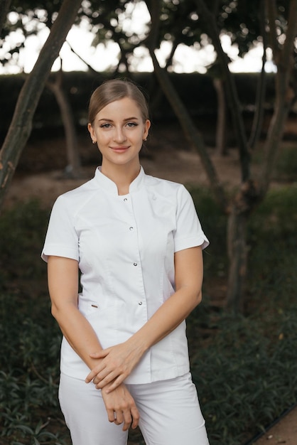 Eine Frau in weißer Uniform steht vor Bäumen und lächelt.