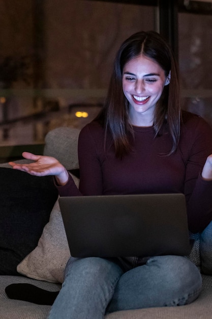 Eine Frau lächelt und amüsiert sich während eines Videogesprächs mit einem Laptop, sitzt bequem zu Hause auf dem Sofa in der dunklen Nacht, Leute plaudern auf einem Notebook mit Internetverbindung