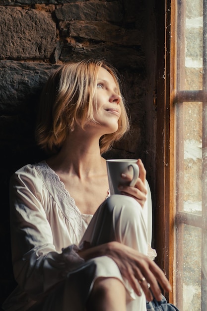 Eine Frau lächelt wehmütig und trinkt morgens Kaffee am Fenster. Frau mittleren Alters mit blonden kurzen Haaren. Morgenentspannung. Weißes Hemd