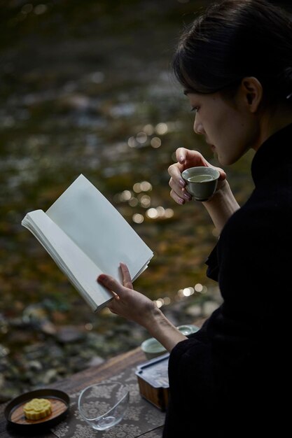 Eine Frau liest am Fluss in den Bergen ein Buch, trinkt Tee und taucht in die Natur ein