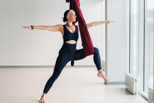 Eine Frau macht Yoga auf einer hängenden burgunderfarbenen Hängematte in einem hellen Fitnessstudio.