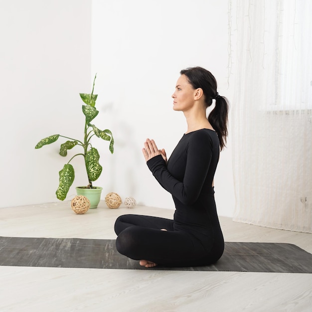 Eine Frau meditiert sitzend in einer Lotusposition mit Namaste-Zügen in schwarzer Sportkleidung