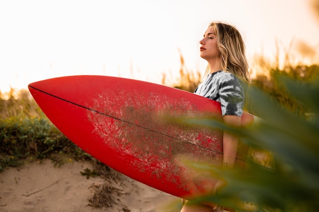 Eine Frau mit blonden Haaren, die im Sommer mit einem Surfbrett auf dem Sand des Strandes spaziert