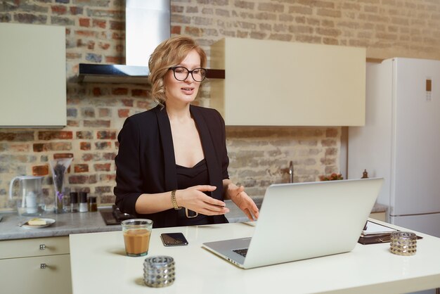 Eine Frau mit Brille arbeitet in ihrer Küche an einem Laptop. Ein gestikulierendes blondes Mädchen diskutiert mit ihren Geschäftspartnern auf einer Videokonferenz zu Hause.