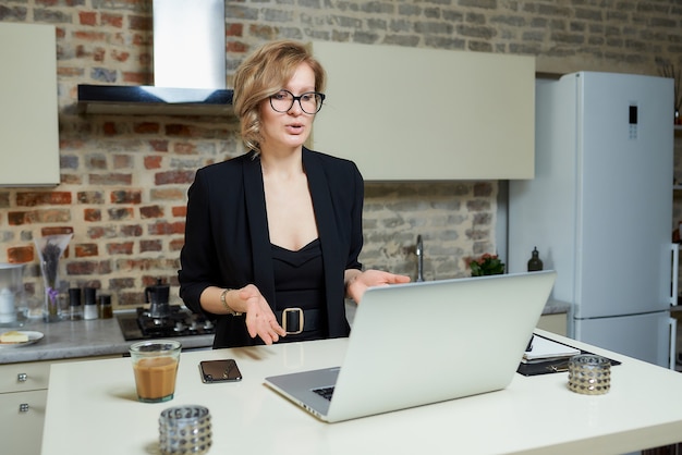 Eine Frau mit Brille arbeitet in ihrer Küche an einem Laptop. Ein gestikulierendes blondes Mädchen diskutiert mit ihren Kollegen über ein Online-Business-Briefing zu Hause.