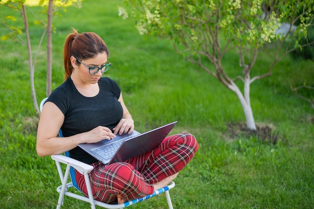 Eine Frau mit Brille sitzt mit gekreuzten Beinen im Lotussitz auf einem Klappstuhl und tippt auf einem Laptop. Eine Freiberuflerin arbeitet während der Quarantäne aus der Ferne im Garten der Hütte