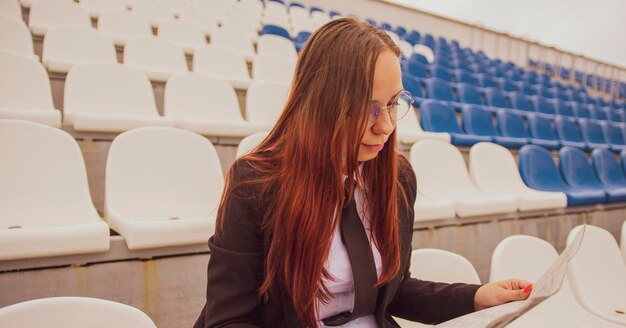 Eine Frau mit Brille und Business-Anzug liest Zeitung, während sie im Stadion sitzt