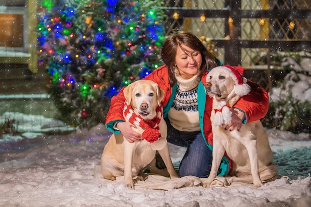 Eine Frau mit einem goldenen Labrador in einem Schal sitzt bei einem Schneefall im Winter im Innenhof eines Wohnhauses neben einem geschmückten Weihnachtsbaum und Schlitten.