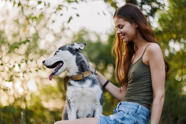 Eine Frau mit einem Husky-Hund lächelt und streichelt liebevoll ihren geliebten Hund, während sie im Herbst vor dem Hintergrund des Sonnenuntergangs in der Natur im Park spazieren geht