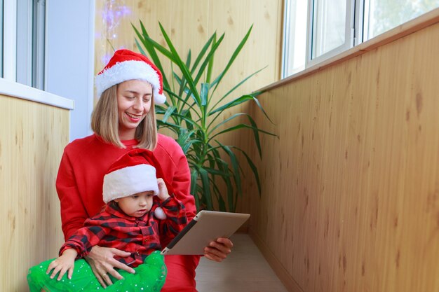 Eine Frau mit einem Kind in roten Weihnachtsmützen gratuliert Familie oder Freunden mit einer Tablette.