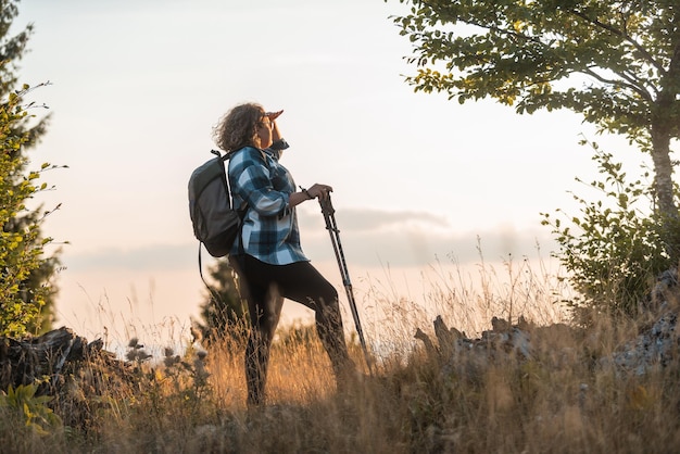 Eine Frau mit einem Rucksack auf dem Rücken und Bergsteigerausrüstung, die bei Sonnenuntergang auf einem Berg spazieren geht