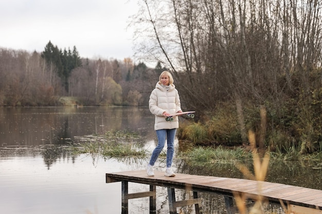 Eine Frau mit einem Skateboard geht in einem Herbstpark am Fluss spazieren und lernt das Skaten am Wochenende