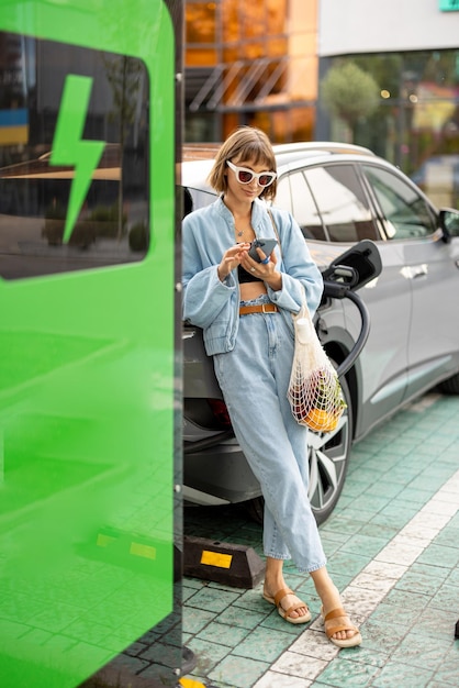 Eine Frau mit einem Smartphone und Lebensmittelwaren, die darauf wartet, dass ihr Elektroauto im Freien geladen wird