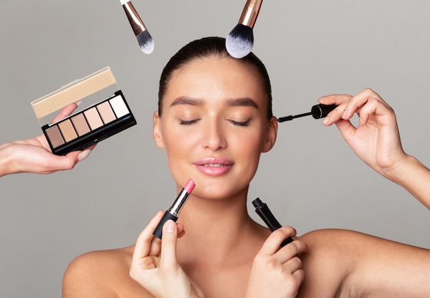Eine Frau mit geschlossenen Augen lässt sich von mehreren Händen verschiedene Make-up-Produkte auf ihr Gesicht auftragen