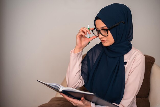 Eine Frau mit Hijab und Brille sitzt mit einem Stift auf einem Stuhl