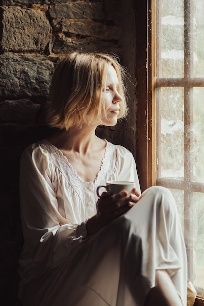 Eine Frau mit nachdenklichem Blick trinkt Kaffee am Fenster. Frau mittleren Alters in einem weißen Hemd. Traurige Gedanken