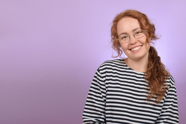 Eine Frau mit roten Haaren und Brille lächelt vor lila Hintergrund.