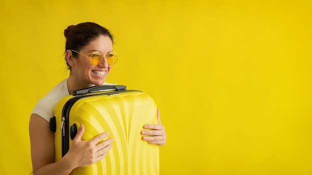 Eine Frau mit Sonnenbrille umarmt einen Koffer auf einem gelben Hintergrund