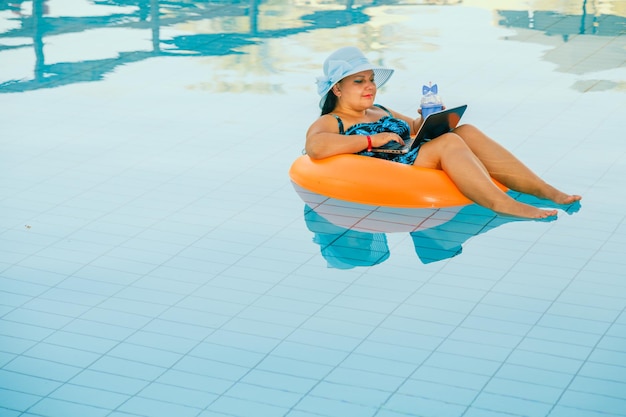 Eine Frau mit Sonnenhut im Pool im Schwimmkreis mit Cocktail und Laptop kommuniziert per Videolink