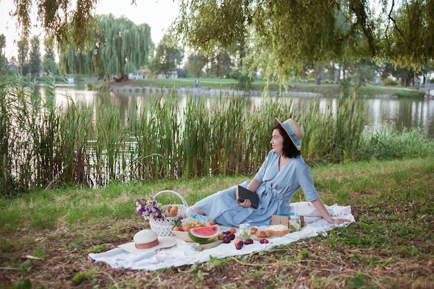 Eine Frau mit Strohhut sitzt bei einem Picknick in einem Park am Ufer eines Flusses