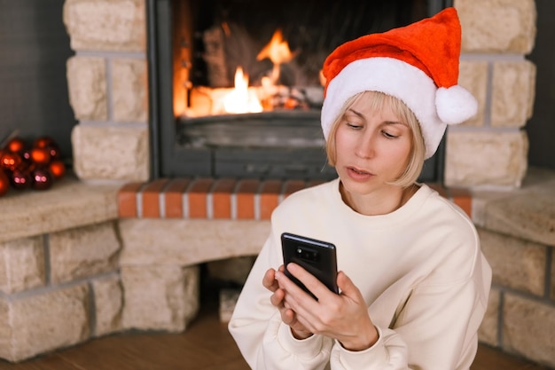 Eine Frau mit Weihnachtsmannmütze kommuniziert am brennenden Kamin in einem gemütlichen Haus am Telefon