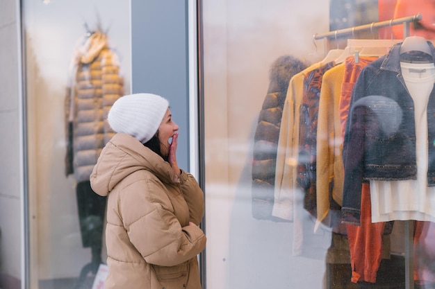 Eine Frau mit weißem Hut und Mantel bedeckt ihren Mund mit der Hand und schaut auf das Schaufenster