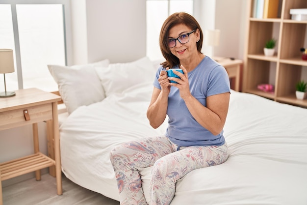 Eine Frau mittleren Alters trinkt eine Tasse Kaffee und sitzt im Schlafzimmer auf dem Bett