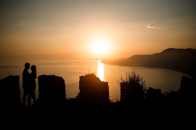 Eine Frau oder ein Mann steht auf einem Felsen und blickt direkt auf den Sonnenuntergang am Meer Das Konzept von Natur und Schönheit Orangefarbener Sonnenuntergang Silhouette bei Sonnenuntergang