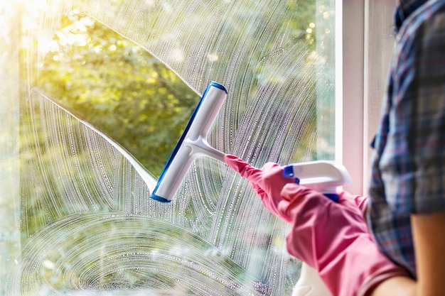 Eine Frau putzt eine Fensterscheibe mit einem Rakel und Seifenlauge. Reinigung mit einem Reinigungsmittel. Hände in rosa Schutzhandschuhen, die Glas an den Fenstern mit einer Sprühflasche, Hausroutine, Hausarbeitskonzept waschen.