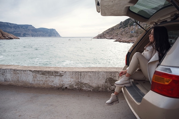 Eine Frau ruht sich im Kofferraum eines Autos aus und blickt aufs Meer. Herbstfahrt bei Sonnenuntergang. Das Konzept der Bewegungsfreiheit. Herbstwochenende. Alleinreisendes oder Alleinreisendes Konzept.