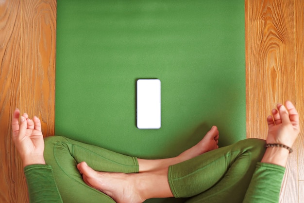 Eine Frau schaut sich Yoga-Kurse auf ihrem Smartphone an, während sie auf einer grünen Gymnastikmatte sitzt. Das Konzept der Ausbildung im Wohnzimmer des Hauses. Technik und Sport