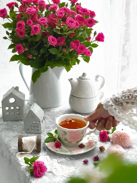 Eine Frau schenkt vor einem Haus mit einem Blumenstrauß eine Tasse Tee ein.