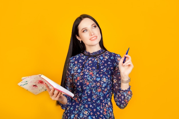 Eine Frau schreibt ihre Ideen auf und hält einen Stift in der Hand und einen Notizblock in der anderen Hand an einer gelben Wand. Konzeptplanung, Geschäftsfrau, Geschäftsidee.