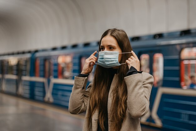 Eine Frau setzt eine medizinische Maske auf ihr Gesicht, um die Ausbreitung des Coronavirus an einer U-Bahnstation zu verhindern. Ein Mädchen mit langen Haaren in einem beigen Mantel wartet auf einem U-Bahnsteig auf einen Zug.