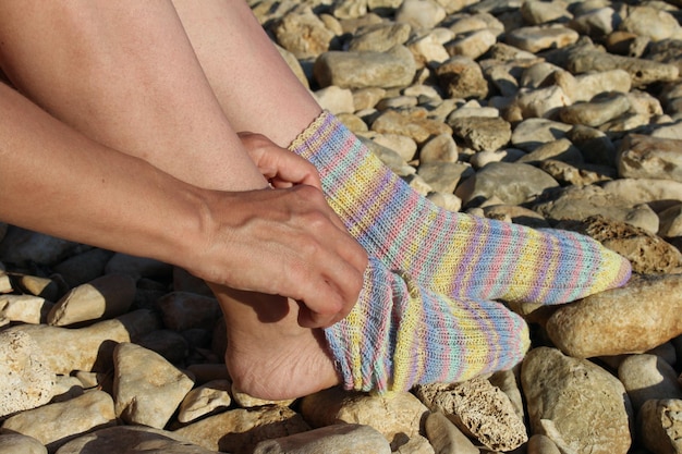 Eine Frau sitzt an einem felsigen Strand, ihre Socken sind zusammengebunden und ihre Füße sind mit einem Streifenmuster bedeckt.