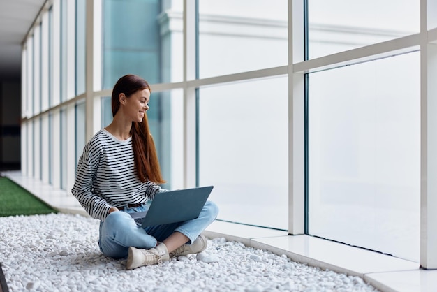 Eine Frau sitzt in der Lobby eines Büros an großen Fenstern und arbeitet an ihrem Laptop online über das Internet. Sie arbeitet freiberuflich überall auf der Welt