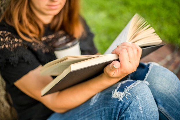 Foto eine frau sitzt in der nähe eines baumes im park und hält ein buch und einen becher mit einem heißen getränk