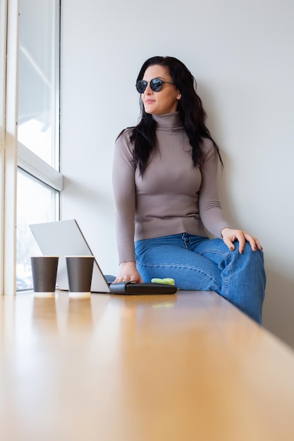 Eine Frau sitzt mit einem Laptop und einer Tasse Kaffee vor einem Fenster.