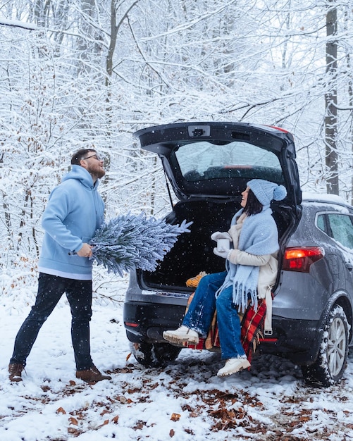 Eine Frau sitzt mit einer Tasse warmem Getränk in einem Winterwald, ein Mann stellt einen Weihnachtsbaum in den Kofferraum