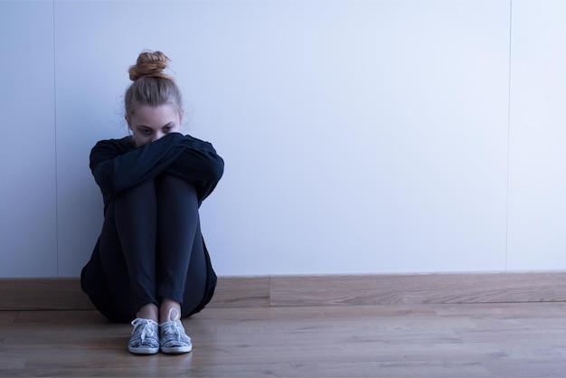 Eine Frau sitzt mit verschränkten Armen und dem Kopf in den Händen auf dem Boden vor einer Wand.