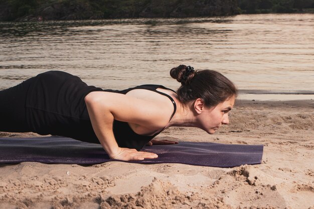 Eine Frau steht in einer Plankenpose am Flussufer am Strand bei Sonnenuntergang im Sommer. Yoga- oder Fitnesskurse in der Natur.