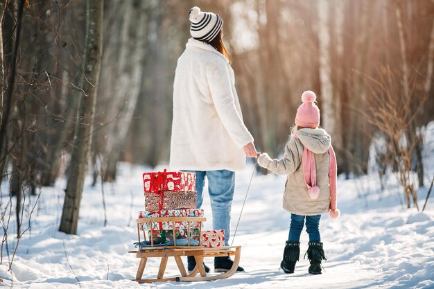 Eine Frau und ein kleines Mädchen tragen den Schlitten mit Weihnachtsgeschenken auf Winterhintergrund