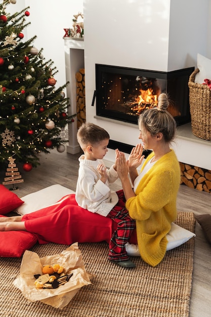 Eine Frau und ihr kleiner Sohn spielen ein Händeklatschspiel neben einem glühenden Kamin in einem gemütlichen Wohnzimmer, das mit einem Weihnachtsbaum und festlicher Dekoration geschmückt ist