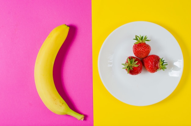 Eine frische Banane und Erdbeeren liegen auf einer gelben und rosa Oberfläche