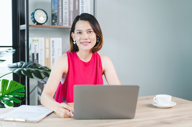 Eine fröhliche asiatische Geschäftsfrau mittleren Alters in entspannter Freizeitkleidung, die zu Hause arbeitet, E-Mails auf Computer-Laptop überprüft und auf Dokumentenpapier der Finanzbuchhaltung schreibt. Geschäft Stockfotos