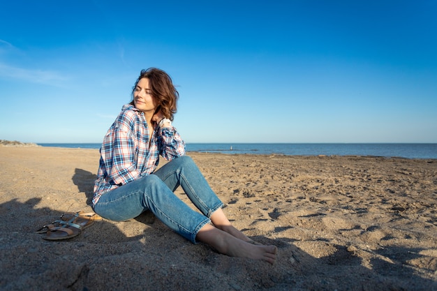 Eine fröhliche dunkelhaarige Frau lächelt, sitzt am Strand und genießt die strahlende Sonne an einem Sommertag. Konzept der Sommerferien auf See und Live-Stil