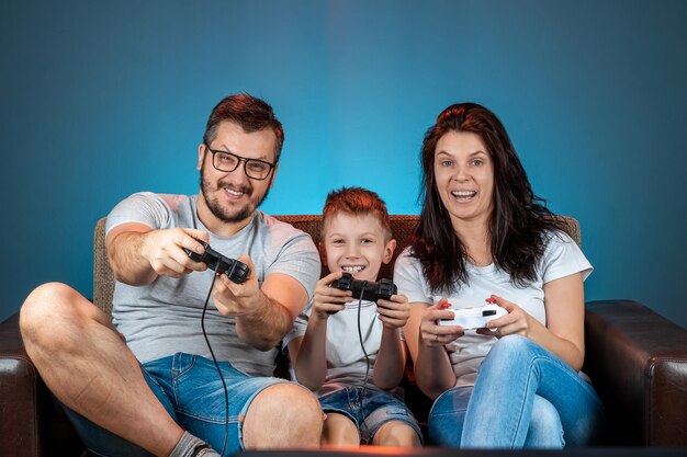 Eine fröhliche Familie, Papa, Mama und Sohn spielen auf der Konsole, Videospiele, reagieren emotional auf der Couch sitzend. Ruhetag, Unterhaltung, Freizeit, Zeit miteinander verbringen.