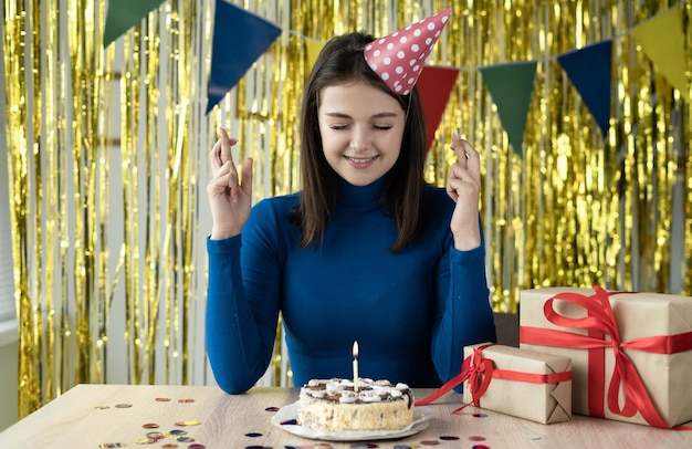 Eine fröhliche Frau mit festlicher Mütze auf dem Kopf sitzt an einem Tisch mit Kuchen und Kerzen und lächelt fröhlich. Daumen drücken macht eine Wunschparty zu Hause online Remote-Geburtstag