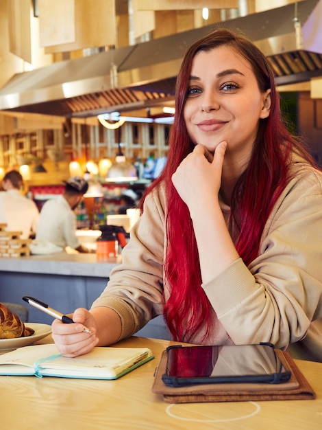 Eine fröhliche Frau mit roten Haaren arbeitet fern in einem Café und schreibt in ein Tagebuch